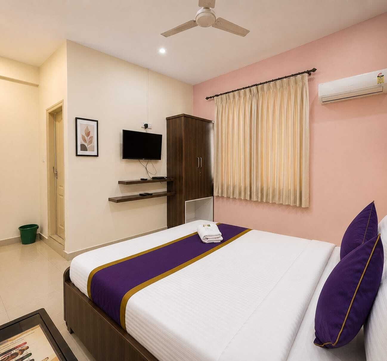 A hotel room with a bed, a television, a wardrobe, a window with curtains, and an air conditioning unit.