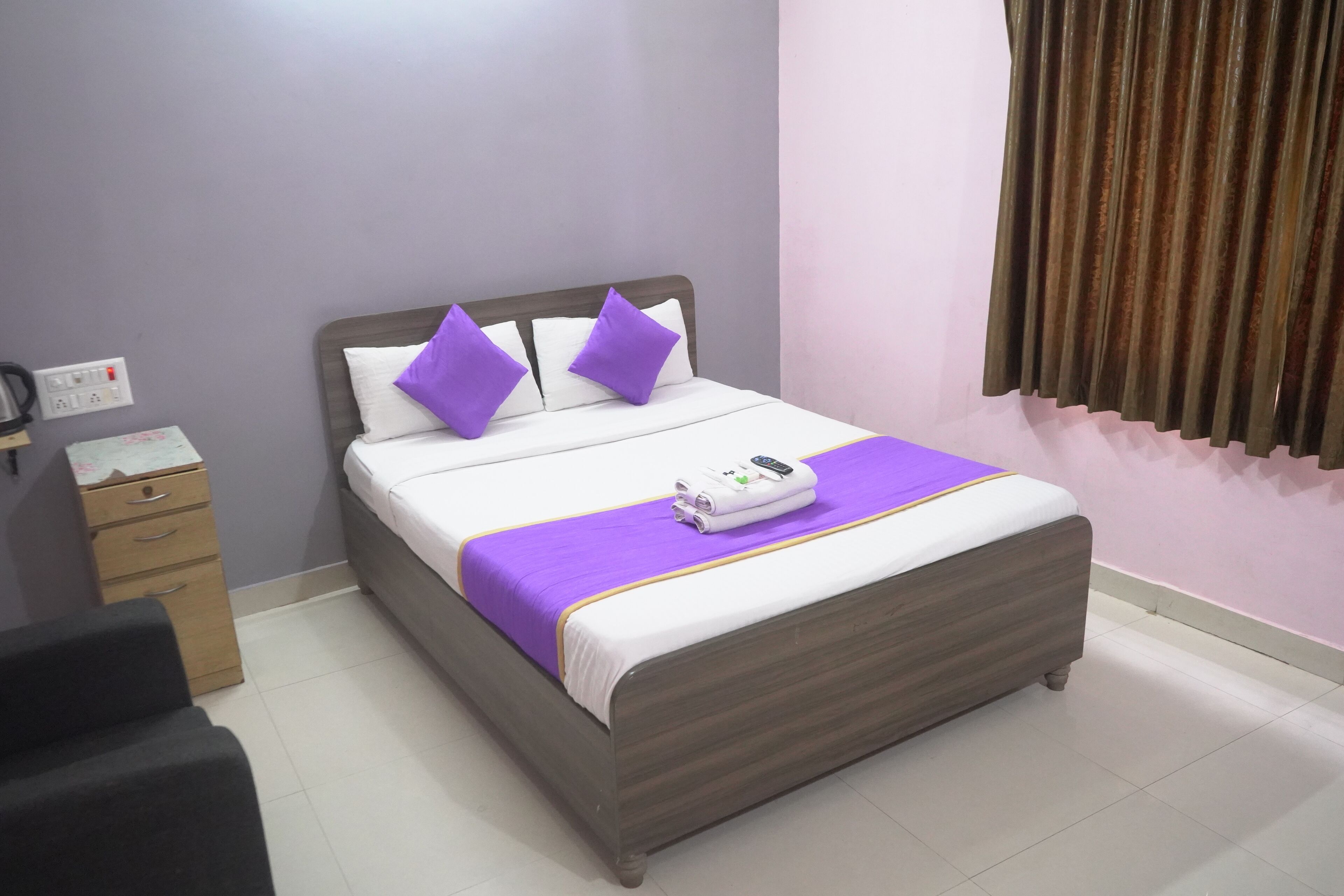 A neatly made bed with a purple blanket and pillows, a bedside table with a kettle, and a power outlet on the wall.