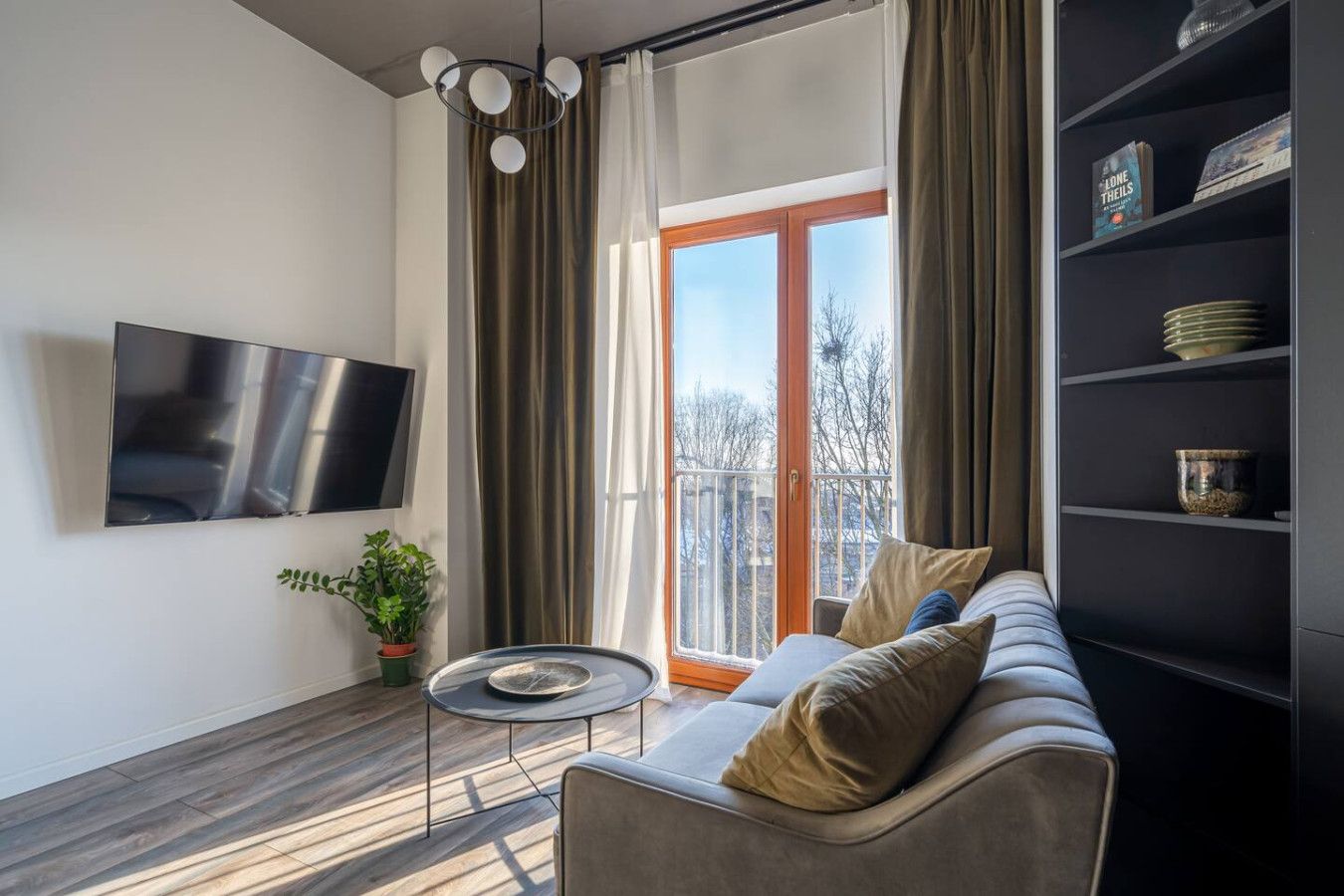 A modern living room with a sofa, a small round table, a flat-screen TV, a bookshelf, and a window with curtains.