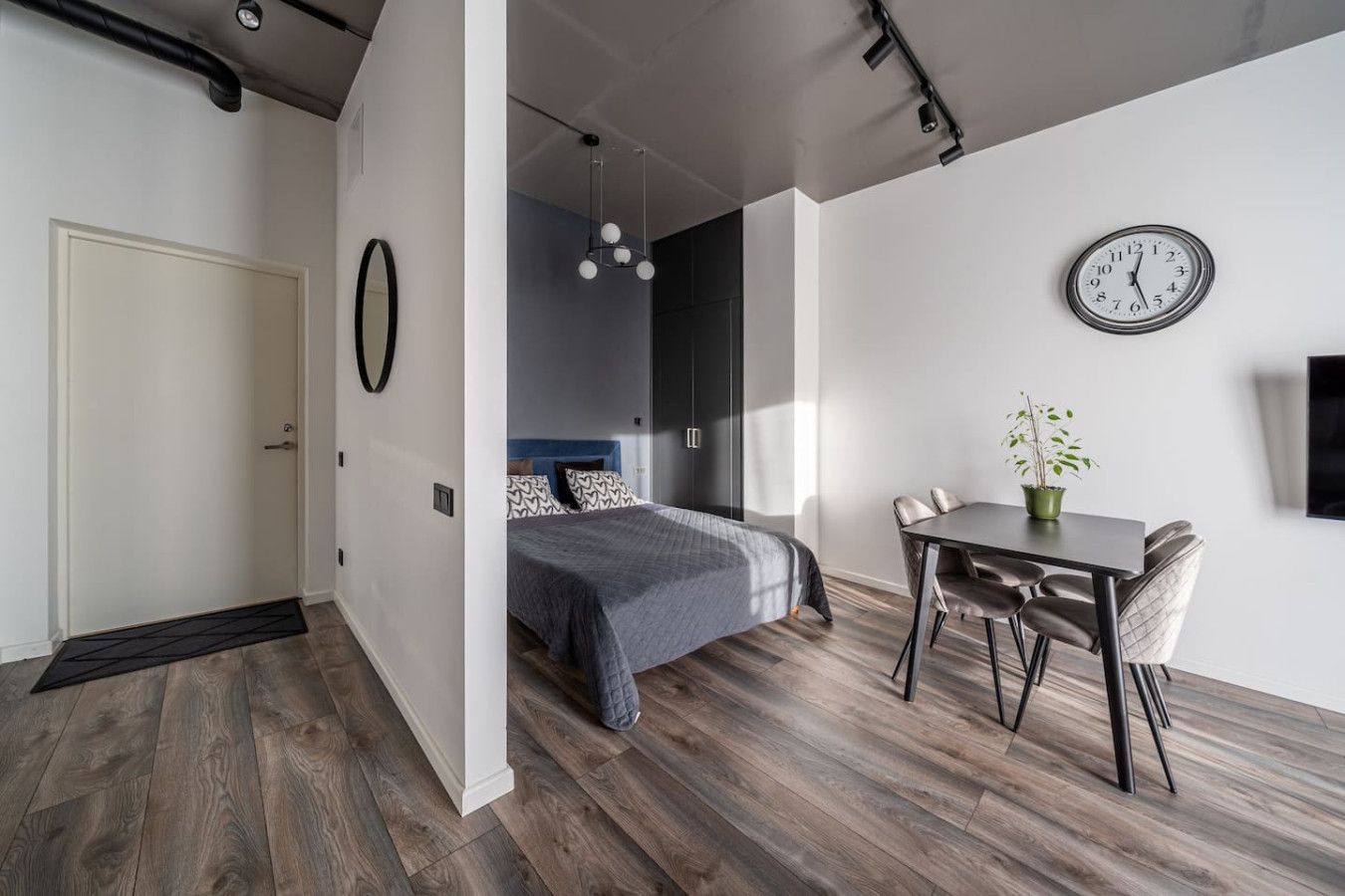 A modern bedroom with a bed, a dining table with chairs, a clock on the wall, and a potted plant.