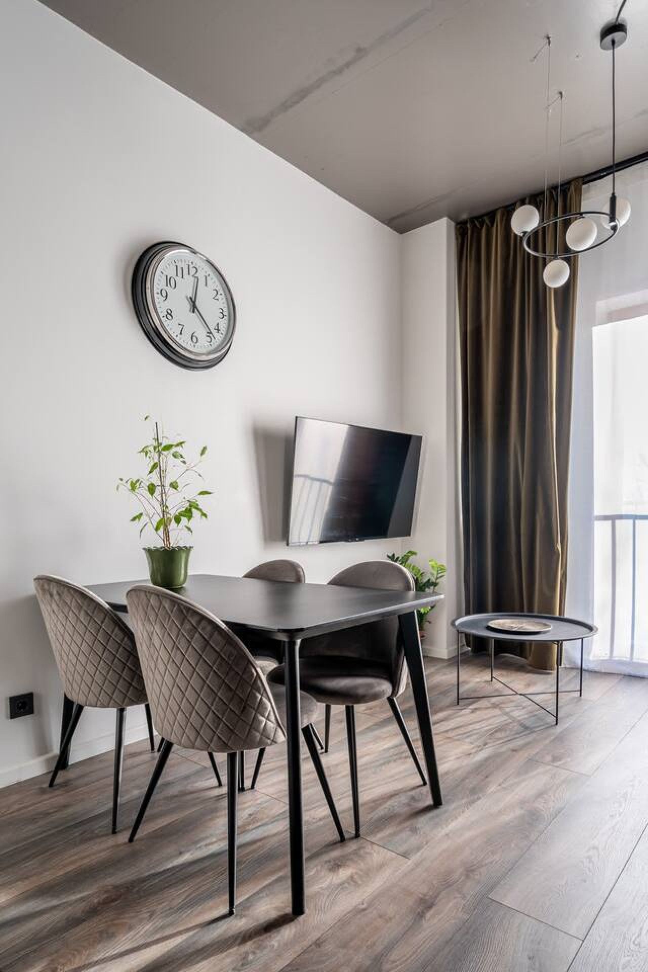 A modern dining area with a wooden table, two chairs, a potted plant, a wall-mounted clock, and a flat-screen TV.