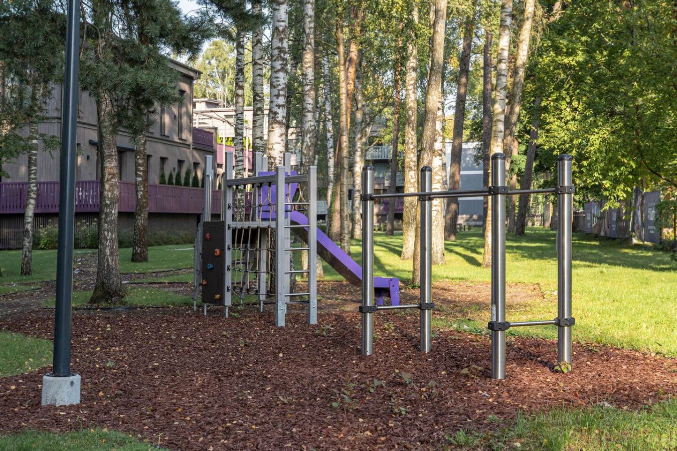 A playground with a slide and climbing structures, surrounded by trees and a building in the background.