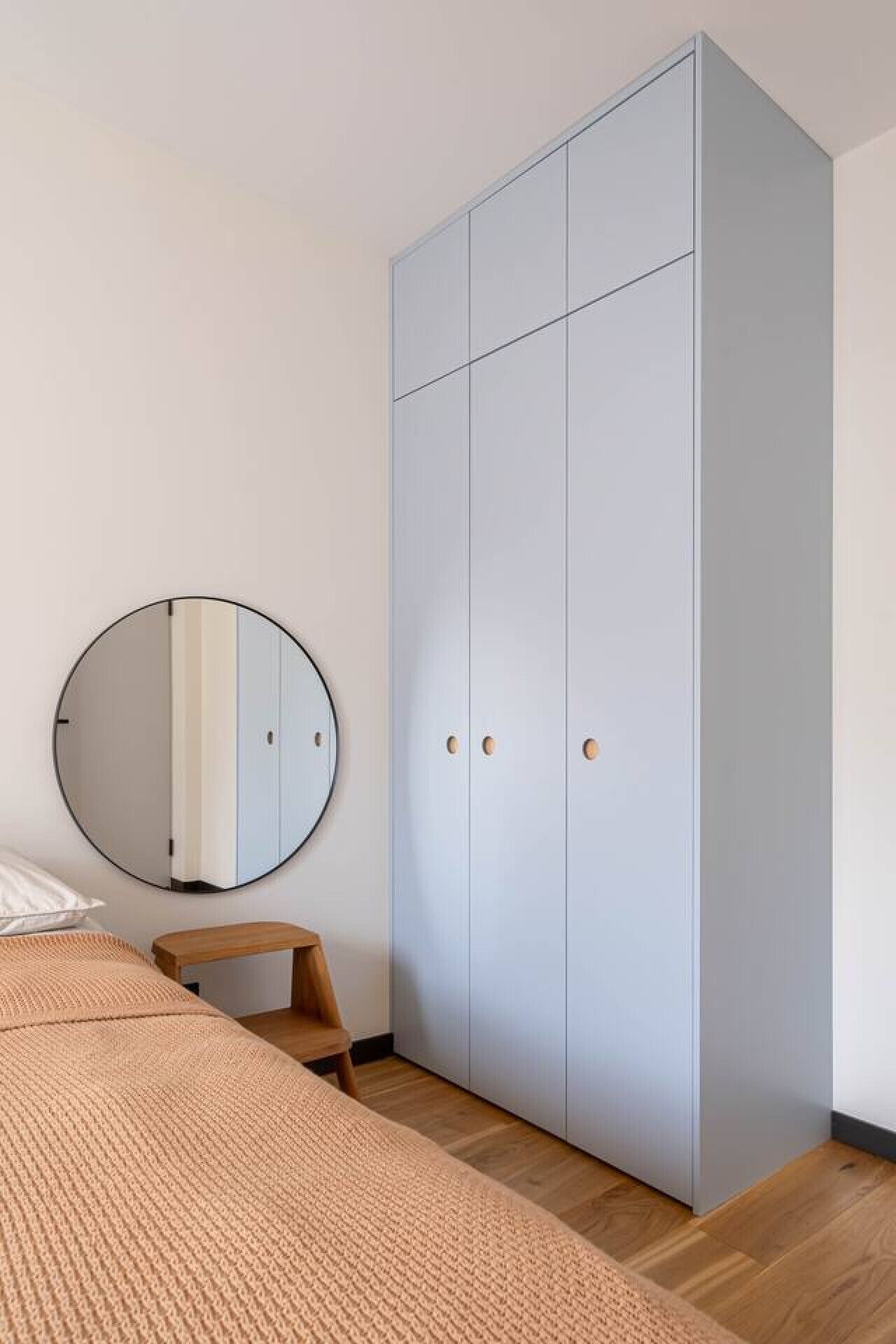 Un dormitorio con un armario grande, un espejo redondo, un banco de madera y una cama con una manta texturizada.