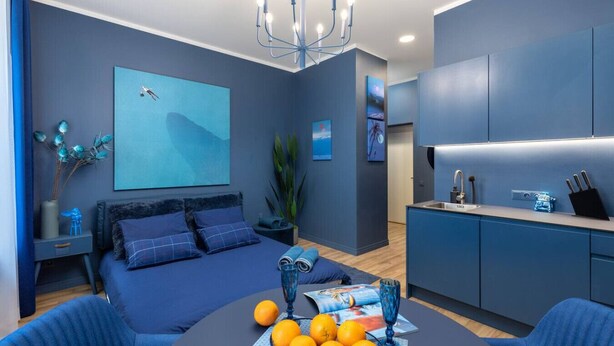 A modern bedroom with a large bed, a bedside table, a nightstand, a desk, a chair, a dining table with oranges and a magazine, a chandelier, and a kitchen area with cabinets and a sink. - Sojourn in Old Town studio apartment (Riga)