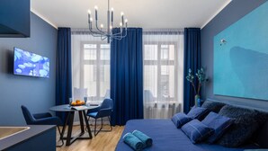 A modern hotel room with a large bed, a desk with a chair, a television, and a view of the city through large windows. - Sojourn in Old Town studio apartment (Riga)