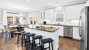 A modern kitchen with white cabinets, a stainless steel refrigerator, and a central island with bar stools. - Sonrise 5-bedroom, 5.5-bathroom home w/ private pool, ~4 mins to the beach! (Ocean Isle Beach)