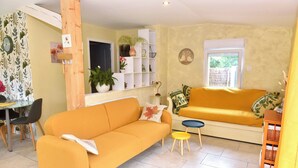 A living room with a yellow sofa, a smaller yellow sofa, a dining table with chairs, and a bookshelf with plants.