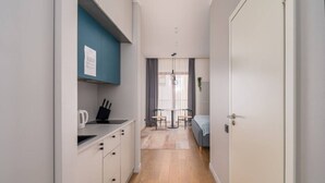 A narrow hallway leading to a dining area with a table and chairs, a kitchenette with a kettle, and a bed with a blue blanket.