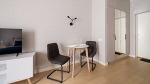 Una sala moderna con un televisor de pantalla plana, dos sillas negras, una mesita redonda con mantel blanco y una lámpara de pared.