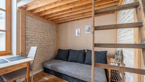 Una habitació acollidora amb sostre de fusta, paret de maó, sofà gris, escriptori de fusta i una cadira.