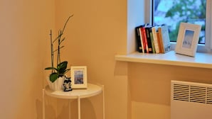 Un coin de pièce avec une petite table blanche sur laquelle sont posés une plante et un tableau encadré, une fenêtre avec des livres et un cadre photo sur le rebord, et un radiateur en dessous.