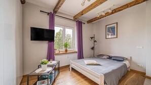 A bedroom with a bed, a TV mounted on the wall, a window with curtains, a bedside table with a plant, and a picture on the wall. - Lacplesa loft-style residence (Riga)