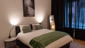 A bedroom with a bed, two bedside lamps, a round side table, a green throw blanket, a black curtain, and a black and white photograph on the wall. - Charming House in Ghent (Gent)
