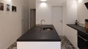 A modern kitchen with a central island, stainless steel sink, and a black countertop. There are three wall-mounted lights above the sink and a door leading to another room. - Charming House in Ghent (Gent)