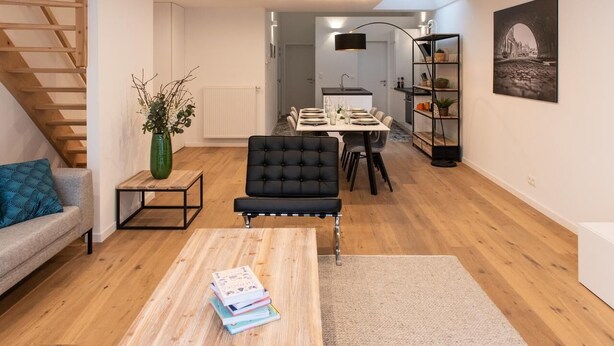 A modern living room with a sofa, a coffee table with books, a dining area with a table and chairs, a staircase, and a bookshelf. - Charming House in Ghent (Gent)