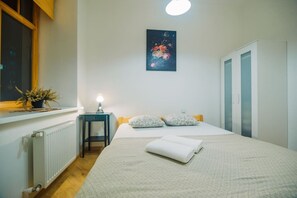 Un dormitorio con cama, mesita de noche, radiador, ventana con planta y un cuadro en la pared.