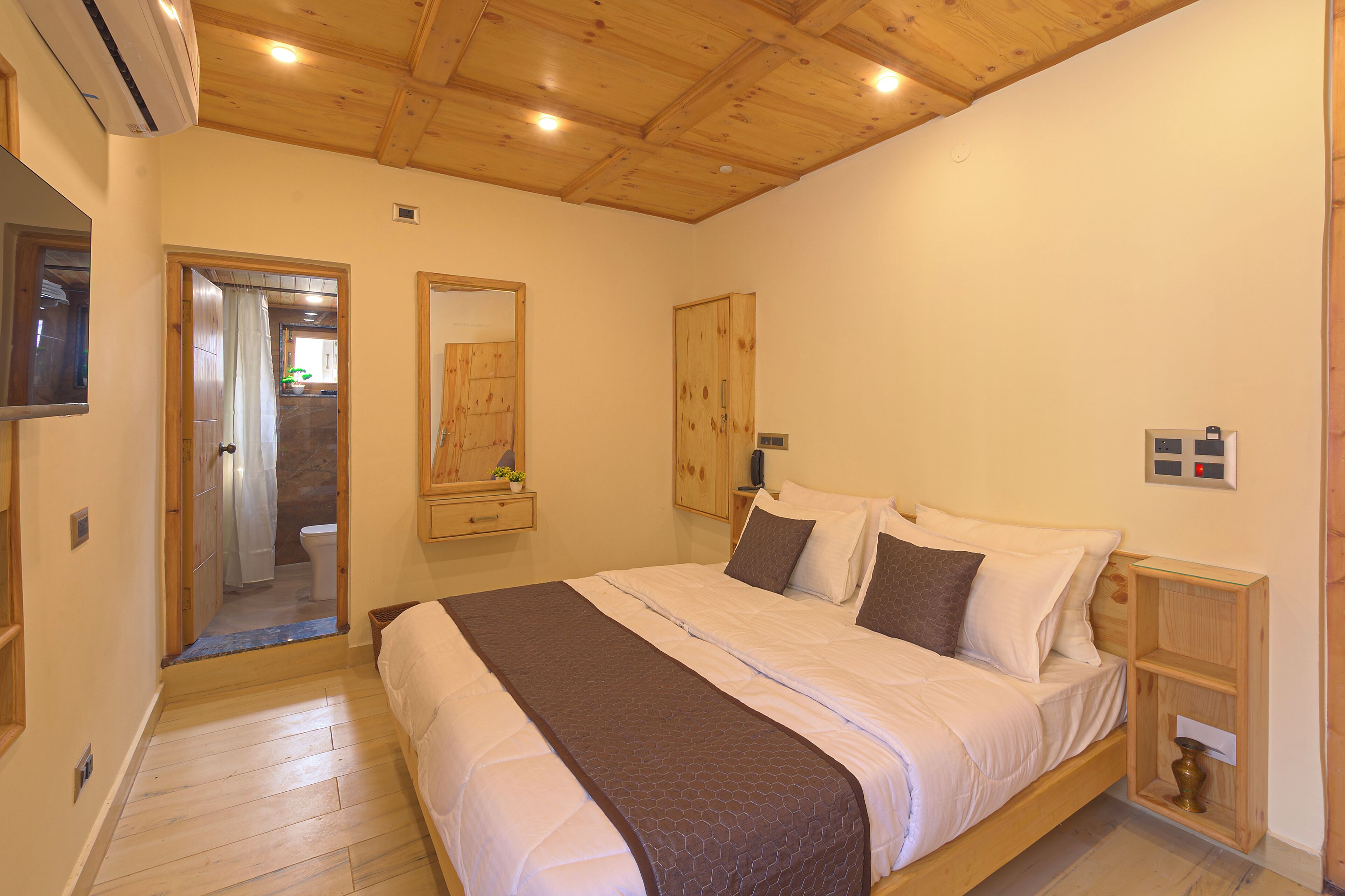 Un dormitorio con cama de madera, una mesita de noche, un espejo y un baño a la vista a través de una puerta abierta.