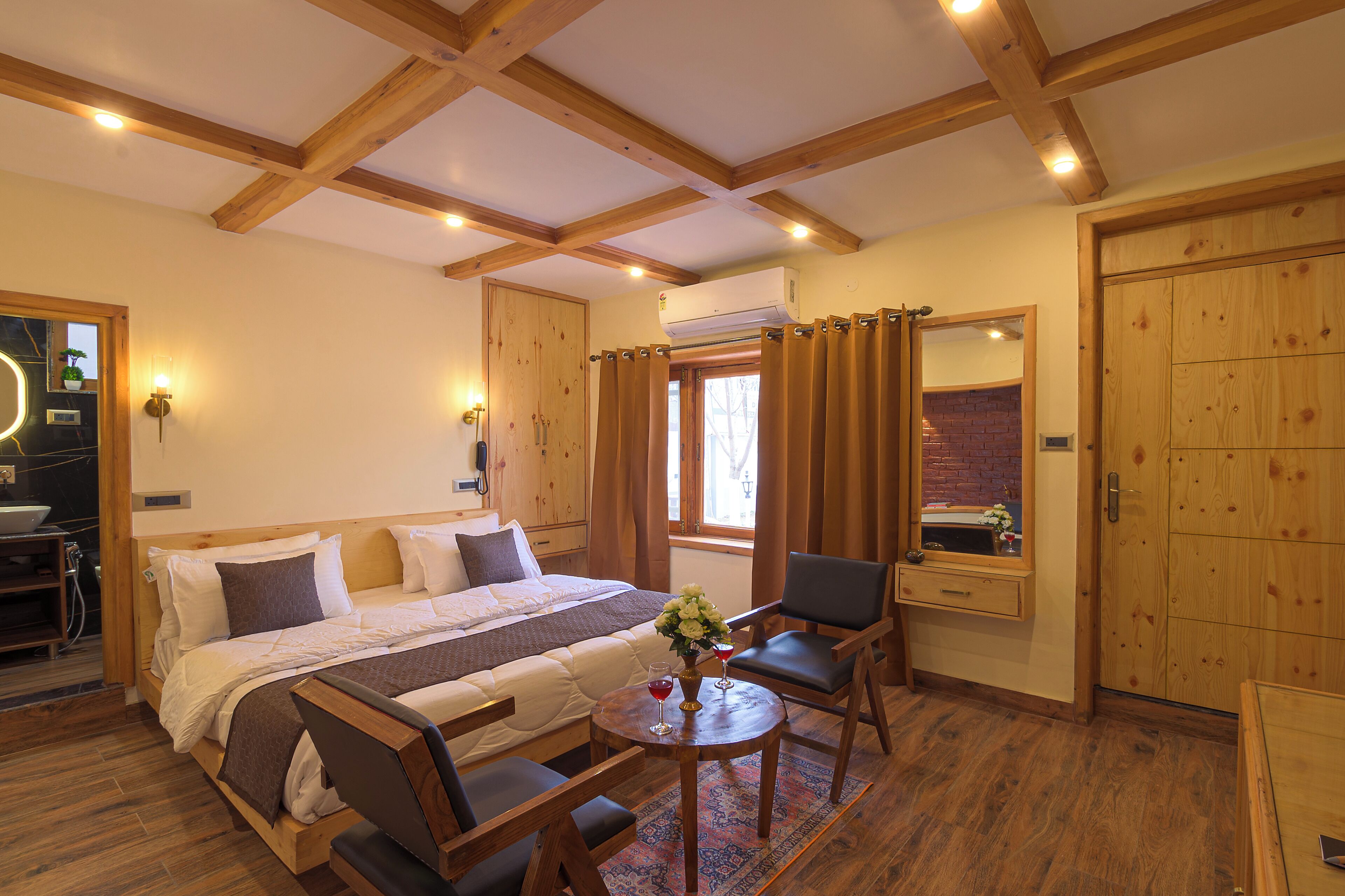 A hotel room with a large bed, wooden ceiling beams, a dining table with chairs, and a small window with curtains.