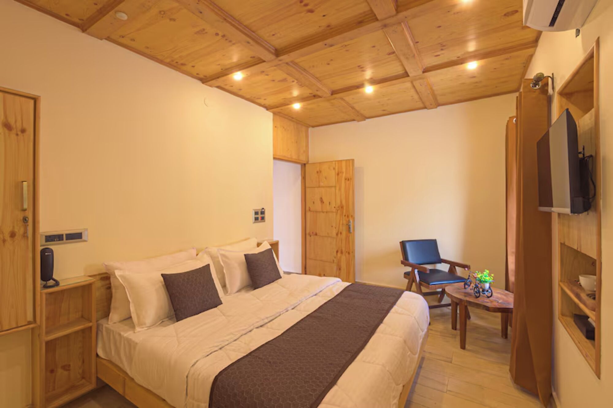 A bedroom with a wooden ceiling, a bed with a brown blanket, a chair, a small table with toys, and a TV on the wall.