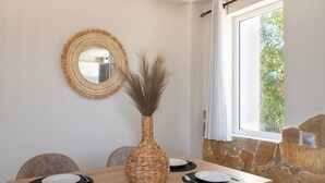 Un coin repas avec une table en bois dressée pour quatre personnes, un vase en osier contenant des plantes séchées, un miroir rond et un mur en pierre.