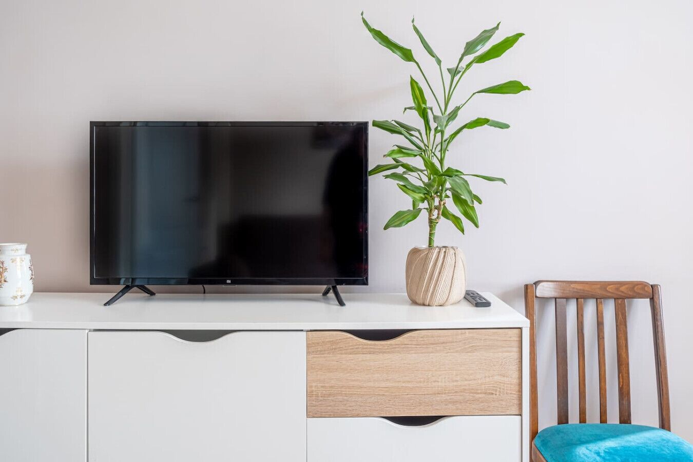 Ein Flachbildfernseher auf einem weißen TV-Schrank, eine Pflanze in einem strukturierten Topf, eine Fernbedienung und ein Holzhocker mit blauer Sitzkissen.