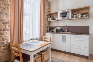 A compact kitchen with a wooden table, chairs, a microwave, a sink, and a shelf with books and kitchenware. - Fresh Studio at Pērnavas 15A | Ground Floor (Riga)
