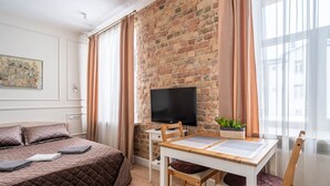 A hotel room with a brick wall, a bed with a quilted cover, a TV on a stand, a dining table with chairs, and a window with curtains.