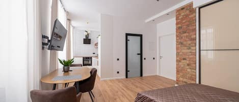 A modern bedroom with a large bed, a dining area with a table and chairs, a kitchen in the background, and a brick accent wall.