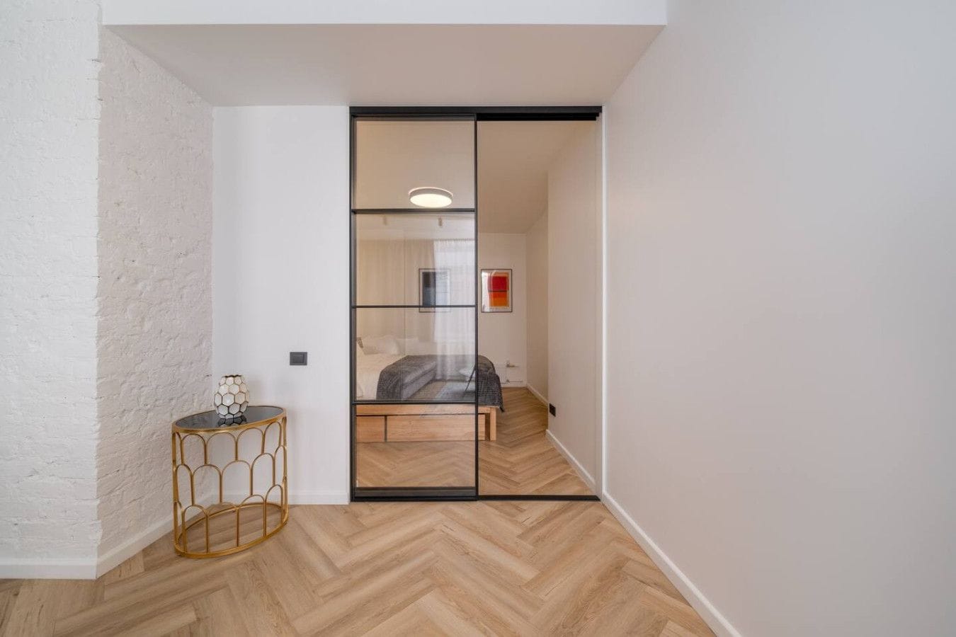 A narrow hallway with a wooden herringbone floor, a glass partition, and a bed with a red accent wall.