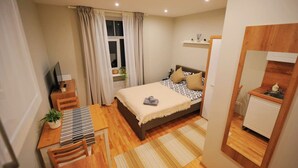A bedroom with a bed, a desk, a chair, a TV, a window with curtains, a small shelf, and a mirror. - Hanzas residence near ArtNouveau quarter & OldTown (Riga)