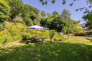 A well-maintained garden with a wooden table and chairs under a blue umbrella, surrounded by lush greenery and trees. - Luxury lodge in peaceful surroundings (saltaash)