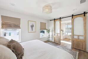 Quarto com uma cama grande, porta deslizante de madeira, janela com persianas, luminária pendente e um quadro na parede.