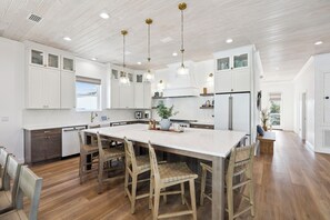A modern kitchen with white cabinetry, a large island, wooden chairs, pendant lights, and a view of the outdoors.