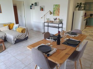 A dining area with a wooden table set for four, a sofa in the background, and a kitchen in the adjacent room. - L' Aparté Nature; gîte for up to 6 people between mountains and countryside (Jarrie)