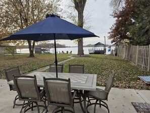 A patio table with six chairs and a blue umbrella, overlooking a lake.