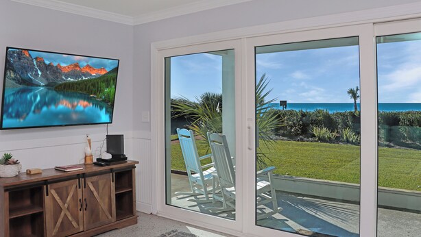 A living room with a large TV, wooden cabinet, and a sliding glass door leading to a patio with chairs and a view of the ocean. - 🌞Steps from the Beach-Pet Friendly-Fab Beach Views-4BR Deja Blue🌞 (Mexico Beach)