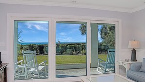 A room with a large sliding glass door leading to a garden, a white rocking chair, a blue chair, and a light blue sofa. - 🌞Steps from the Beach-Pet Friendly-Fab Beach Views-4BR Deja Blue🌞 (Mexico Beach)