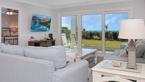 A modern living room with a sofa, a glass coffee table, and a view of the outdoors through large sliding doors. - 🌞Steps from the Beach-Pet Friendly-Fab Beach Views-4BR Deja Blue🌞 (Mexico Beach)