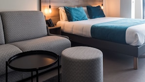 A modern hotel room with a bed, a sofa, a round table, and a small ottoman. - Hôtel des Halles (Dijon)