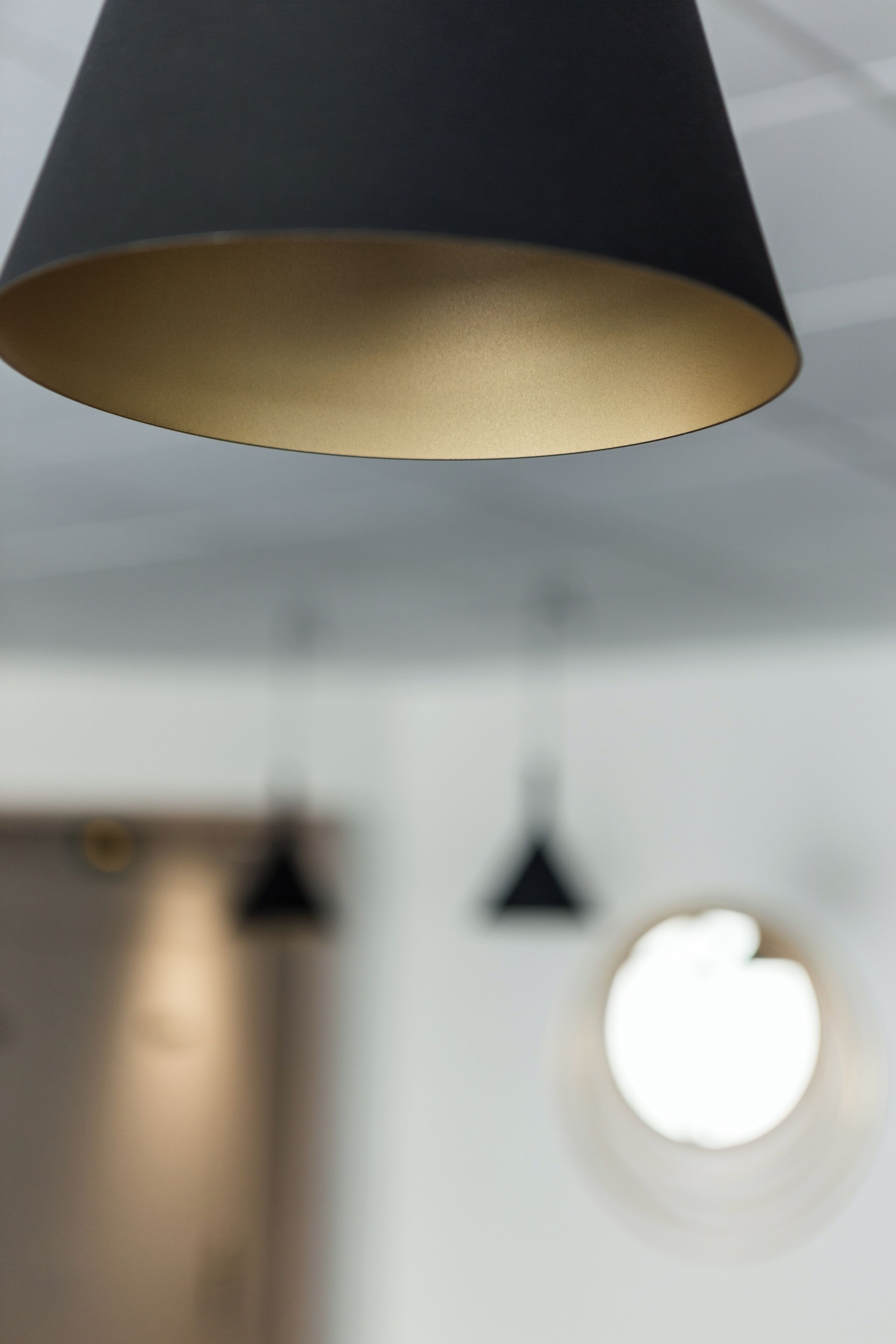 A modern pendant light with a dark shade and a gold-colored base, hanging in a room with a blurred background.