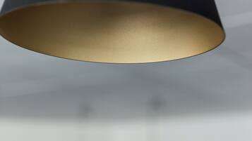 A modern pendant light with a dark shade and a gold-colored base, hanging in a room with a blurred background.