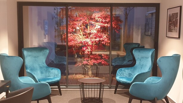 A modern interior with two blue armchairs, a small round table, and a large glass window with a view of a red-leafed tree. - Hôtel des Halles (Dijon)