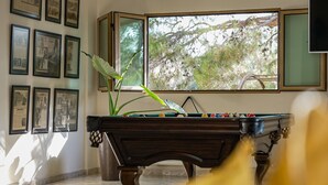 A billiards table in a room with framed pictures on the wall and a window with a view of trees.
