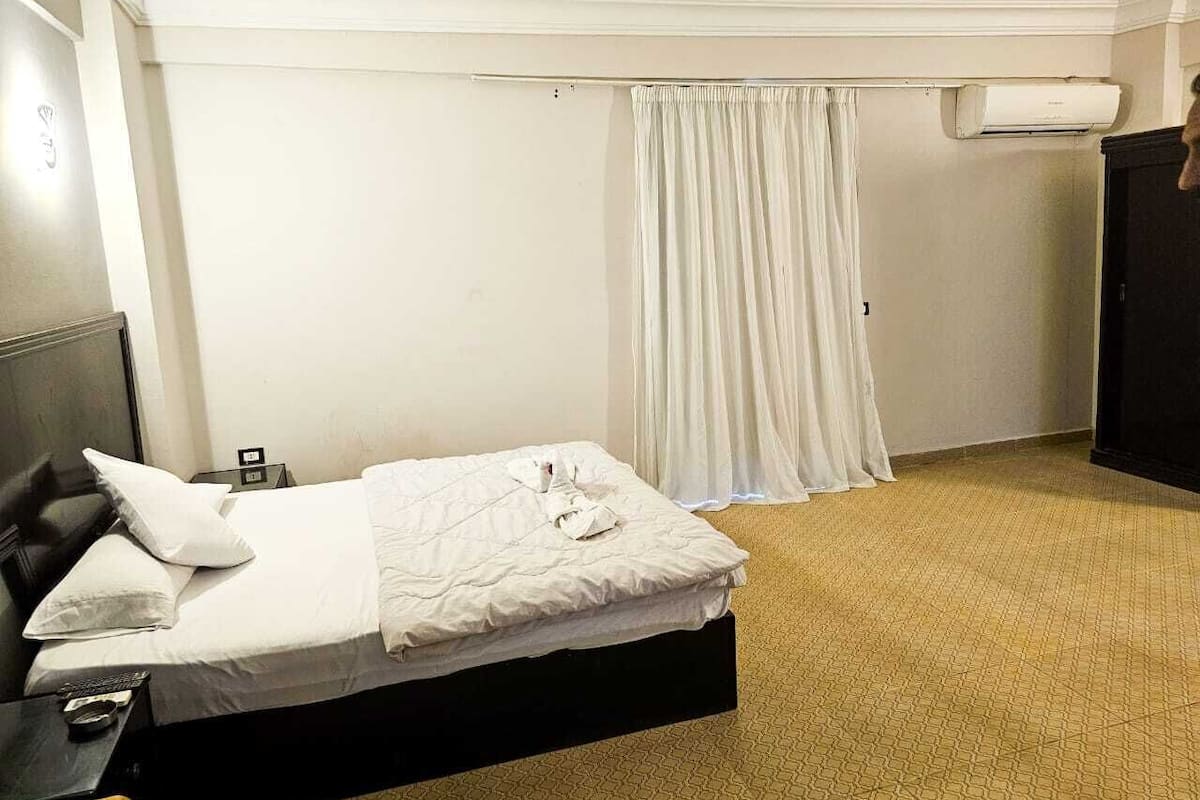 A hotel room with a bed, a nightstand, a desk, a chair, a wardrobe, and an air conditioning unit.
