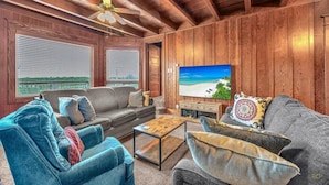 Un salon chaleureux avec des murs en bois, un ventilateur de plafond, un grand téléviseur à écran plat et une vue sur la plage par les fenêtres.