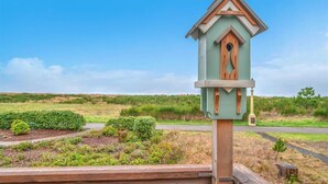 Une nichoir en bois, posé sur un poteau et surmonté d’un toit vert, se détachant sur un paysage composé d’un champ verdoyant et d’un ciel dégagé.