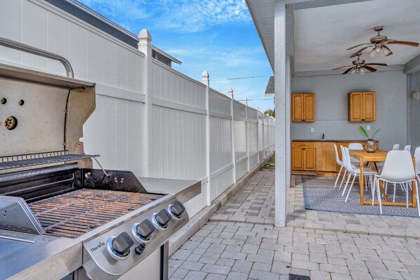 Patio trasero con una parrilla, una mesa de comedor con sillas y una zona de cocina con gabinetes de madera.