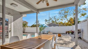 A covered patio area with a wooden table and chairs, a pool, and a white fence in the background.