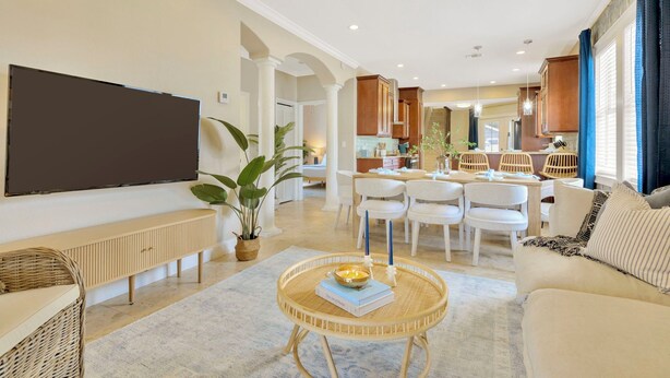 A modern living room with a flat-screen TV, a round coffee table with books and candles, a sofa, and a dining area with white chairs and a wooden table.