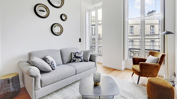 A modern living room with a grey sofa, a round coffee table, and a brown armchair. There are three wall-mounted mirrors and a large window with a view of buildings. - Maison Labruyère (Bordeaux)